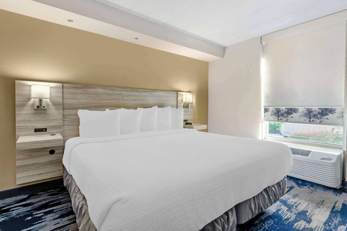 A hotel room with a large bed, a headboard with integrated lighting, a bedside table with a lamp, and a window with a view of trees and a building.