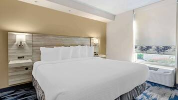 A hotel room with a large bed, a headboard with integrated lighting, a bedside table with a lamp, and a window with a view of trees and a building.