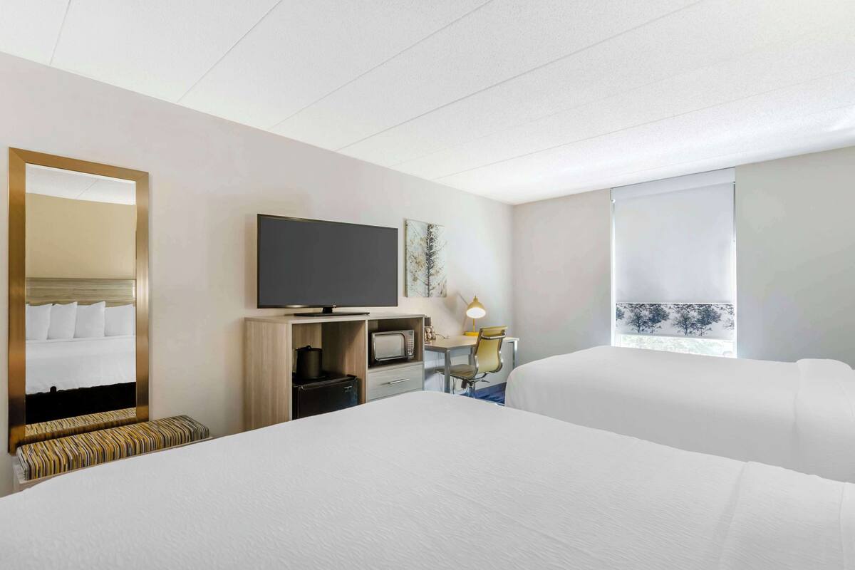 A hotel room with two beds, a television, a desk with a computer, a microwave, and a window with a view of trees.