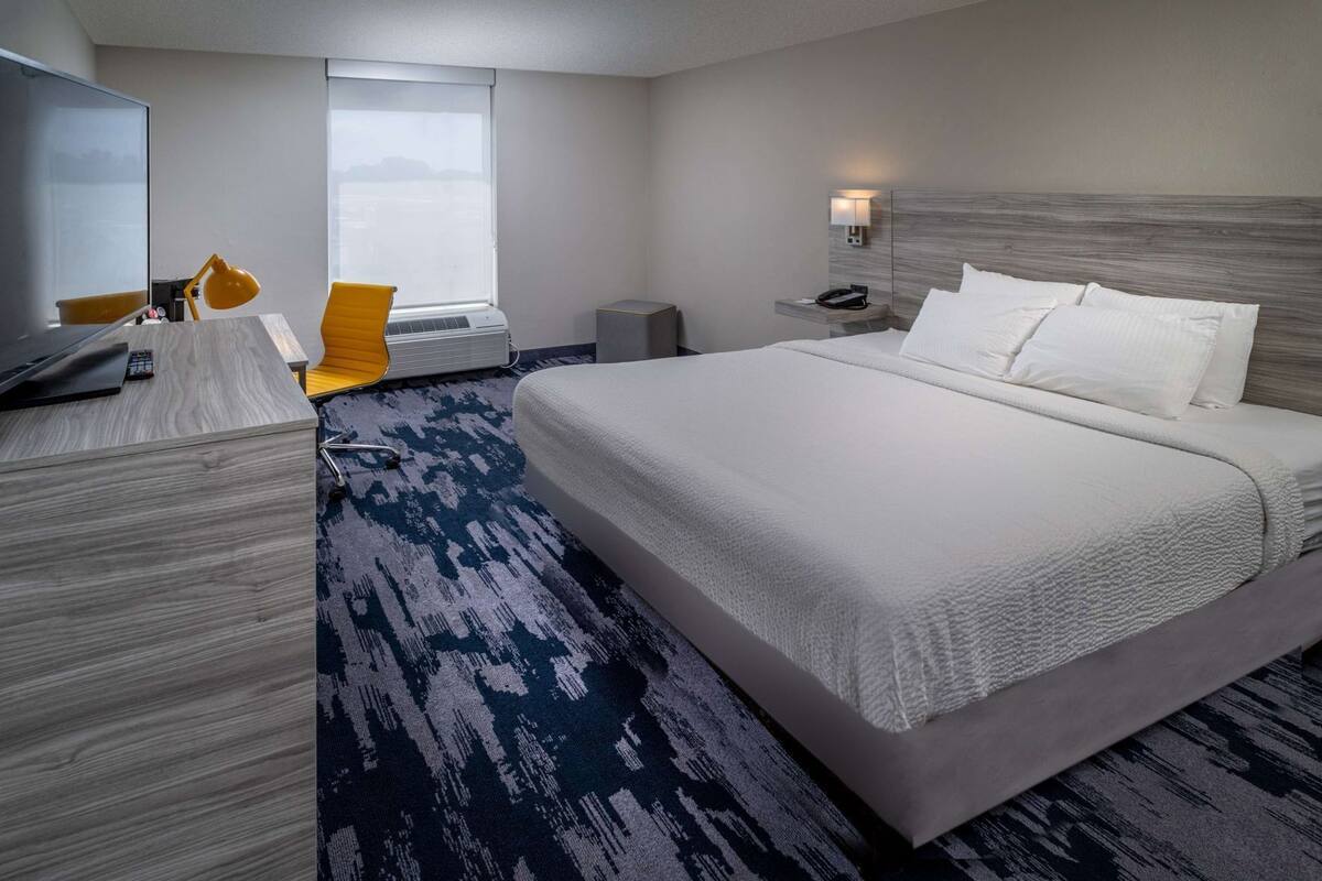 A hotel room with a large bed, a desk with a chair, a desk lamp, a phone, and a window with a view.