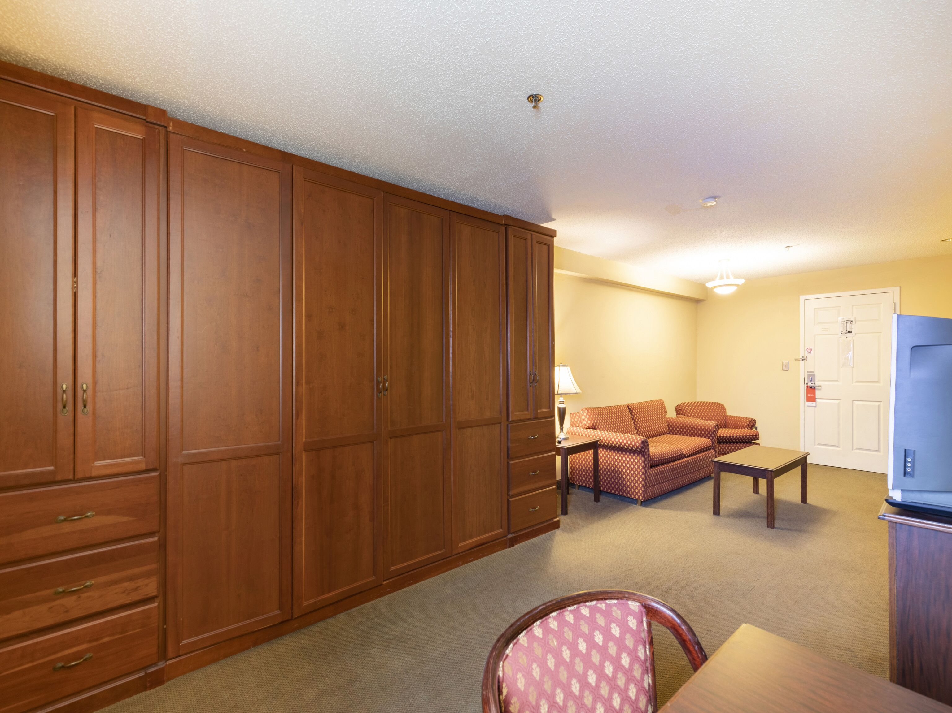 A room with a large wooden wardrobe, a sofa set, a dining table, and a television.