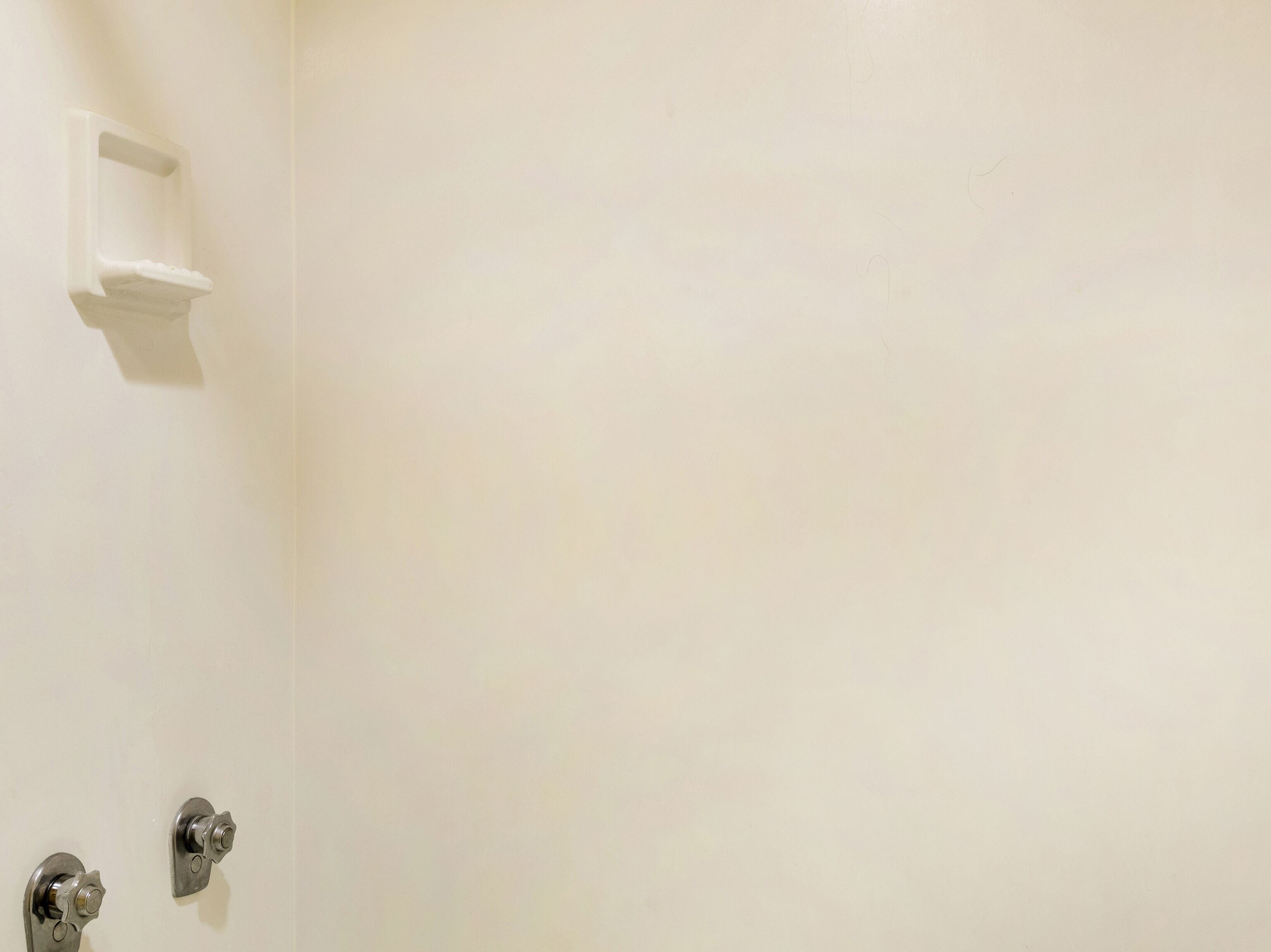 A bathroom with a white wall, a small shelf, and two metallic fixtures.