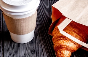 A coffee cup with a lid and a paper bag containing a croissant on a wooden surface.