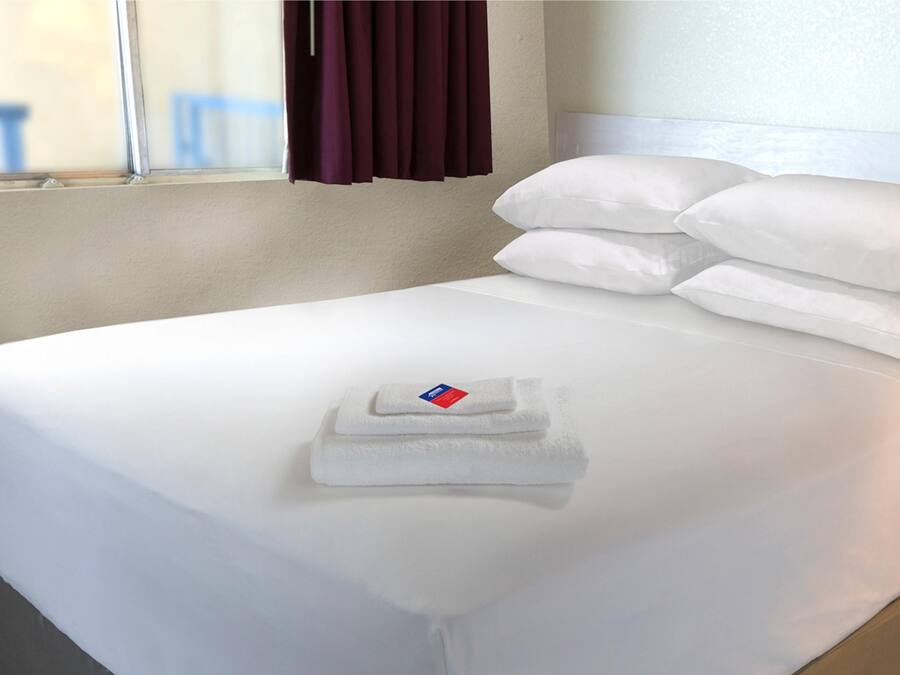 Un lit bien fait, avec du linge de lit blanc et des oreillers, une serviette pliée, et une fenêtre avec des rideaux.