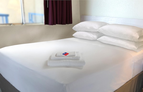 A neatly made bed with white linens and pillows, a folded towel, and a window with curtains.
