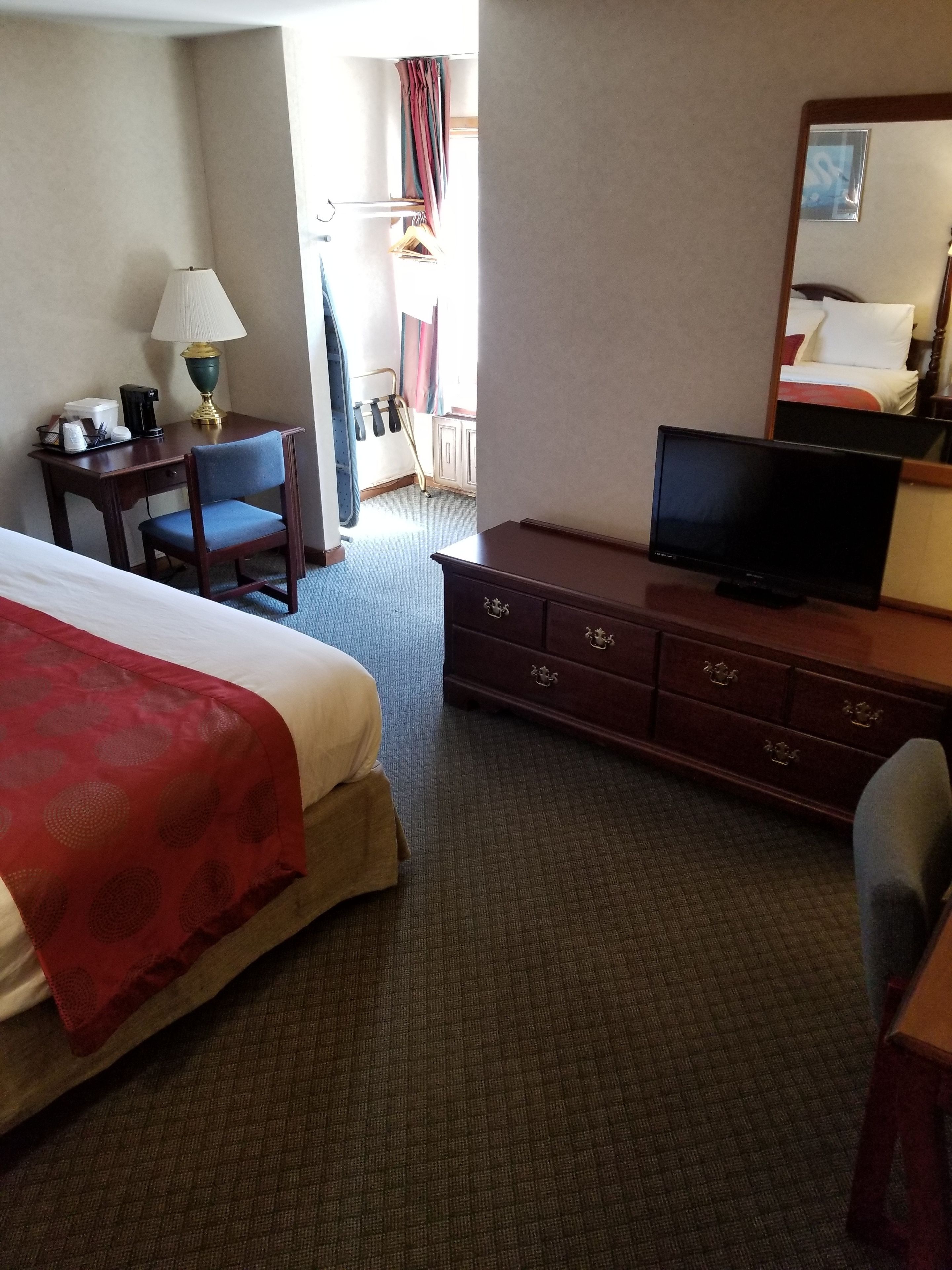 A hotel room with a large bed, a desk with a lamp, a television, and a dresser.