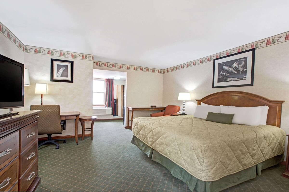Quarto de hotel com cama, escrivaninha, cadeira, televisão e vista para o corredor.