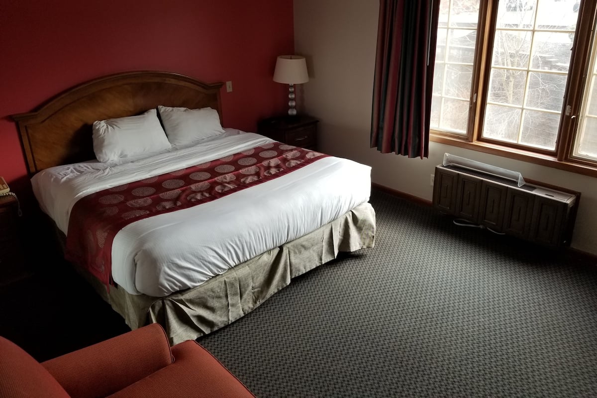 Quarto de hotel com uma cama grande, mesa de cabeceira, luminária, radiador e janela com cortinas.