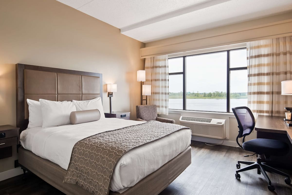 Une chambre d’hôtel avec un grand lit, deux tables de chevet avec des lampes, un bureau avec une chaise et une grande fenêtre avec des rideaux.