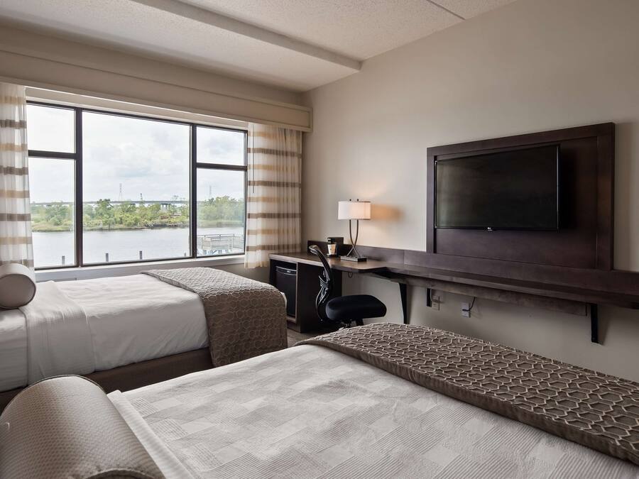 Een hotelkamer met twee bedden, een bureau, een televisie en een raam met uitzicht op een brug en groen.