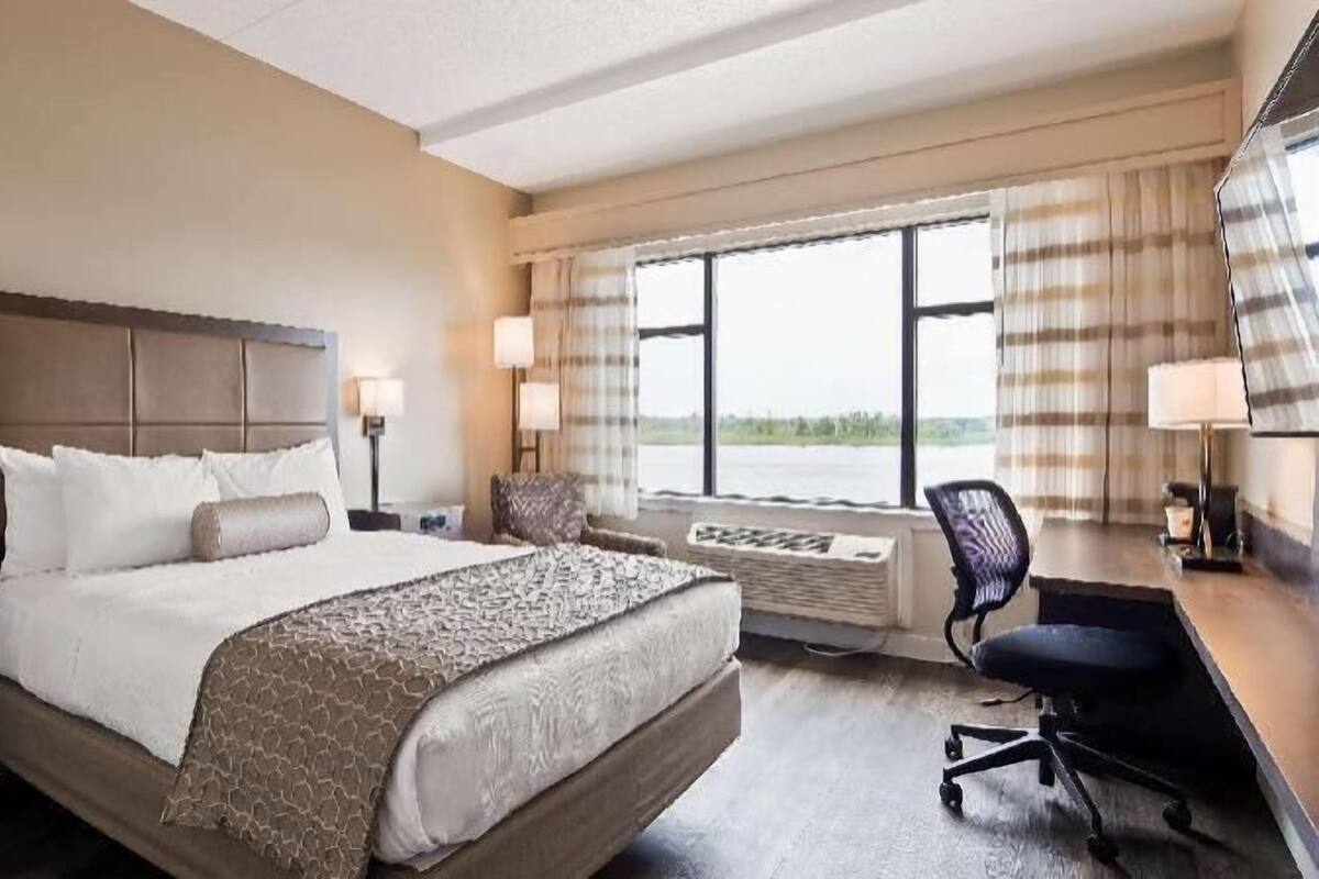 Une chambre d’hôtel avec un grand lit, deux tables de chevet avec des lampes, un bureau avec une chaise et une grande fenêtre avec des rideaux.