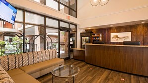 Een hotel lobby met een receptiebalie, een zithoek met een bank en een glazen tafel, en een groot raam met uitzicht op een gebouw van baksteen.