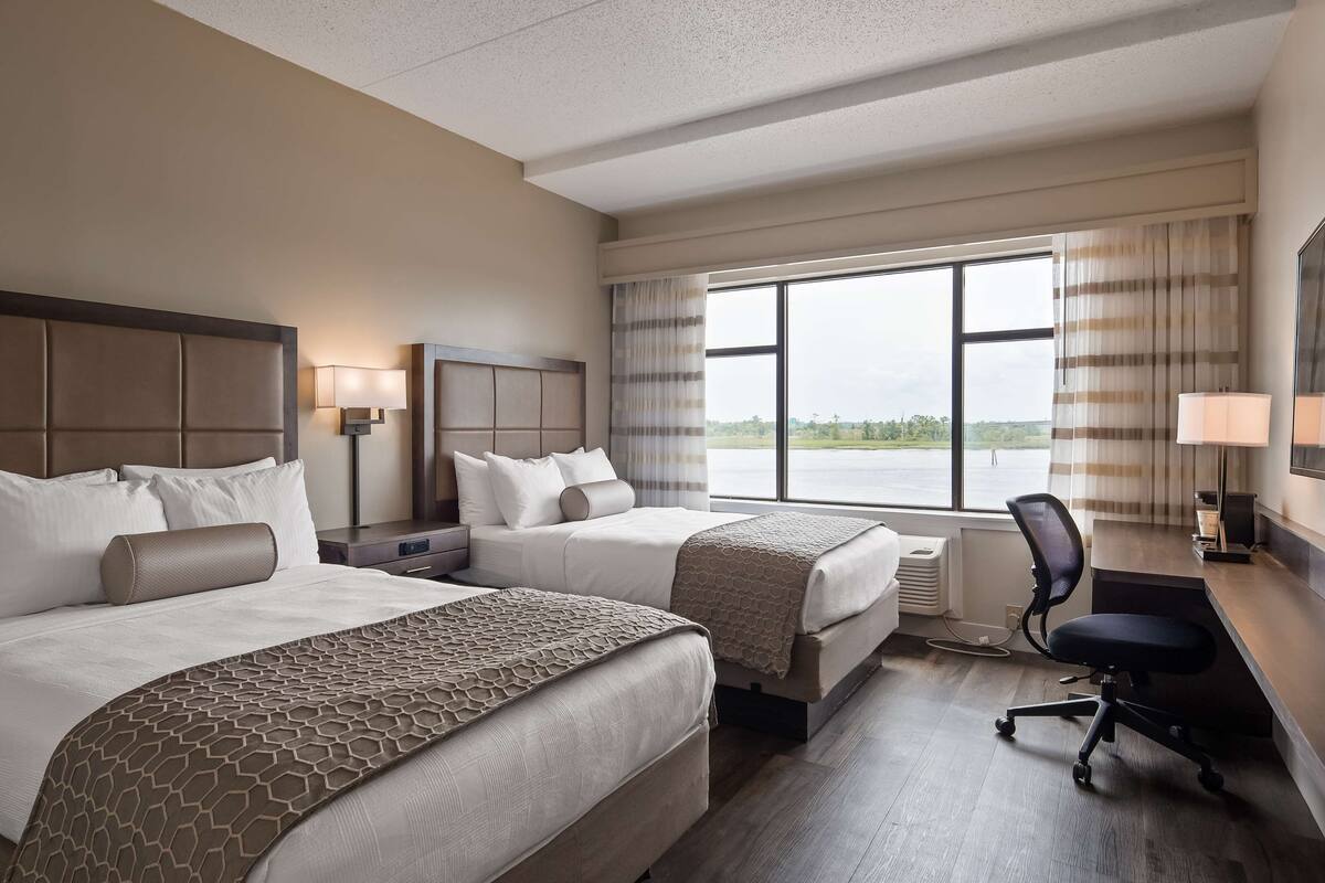 Une chambre d’hôtel avec deux lits, un bureau, une chaise et une grande fenêtre donnant sur un plan d’eau.
