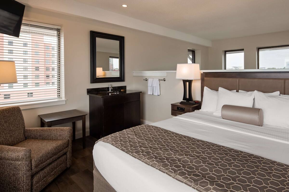 Une chambre d’hôtel avec un grand lit, un bureau, une chaise, une commode, un miroir et une fenêtre avec des stores.