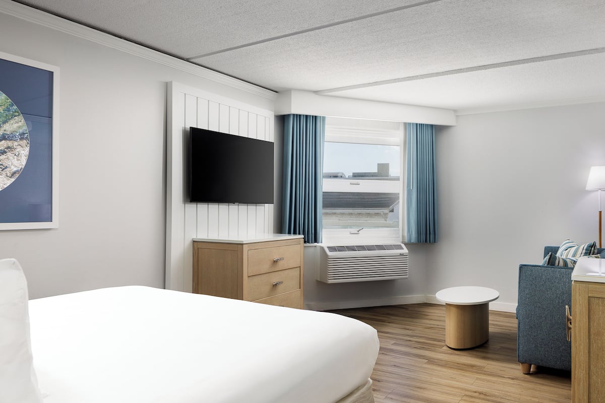 Ein Hotelzimmer mit Bett, Fernseher, Schreibtisch, Stuhl, einem Fenster mit Vorhängen und Blick auf die Stadt.