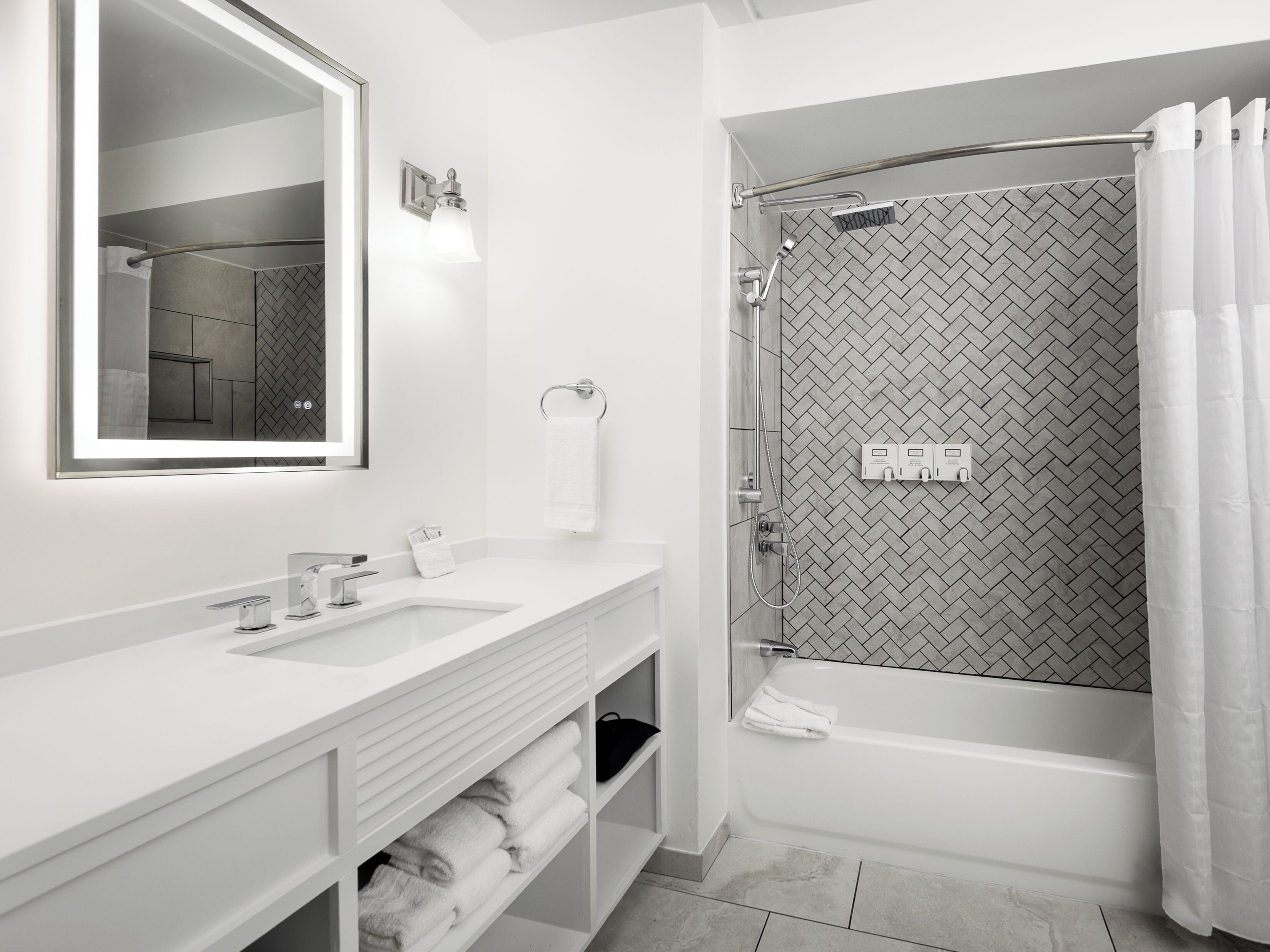 A modern bathroom with a large mirror, two sinks, a shower with a herringbone tile pattern, and a bathtub.