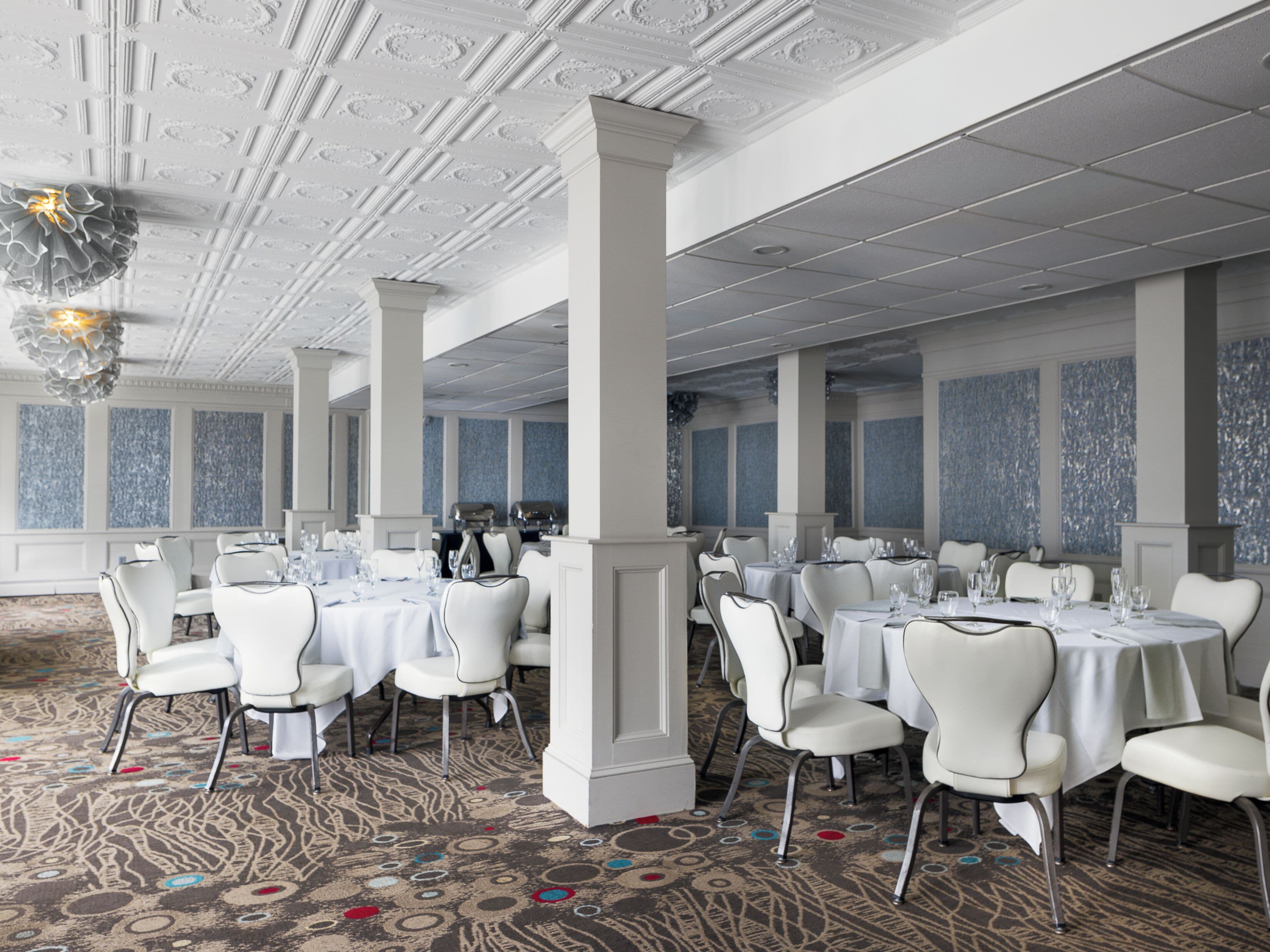 A spacious banquet hall with high ceilings, ornate columns, and a patterned carpet. Tables are set with white tablecloths and chairs.