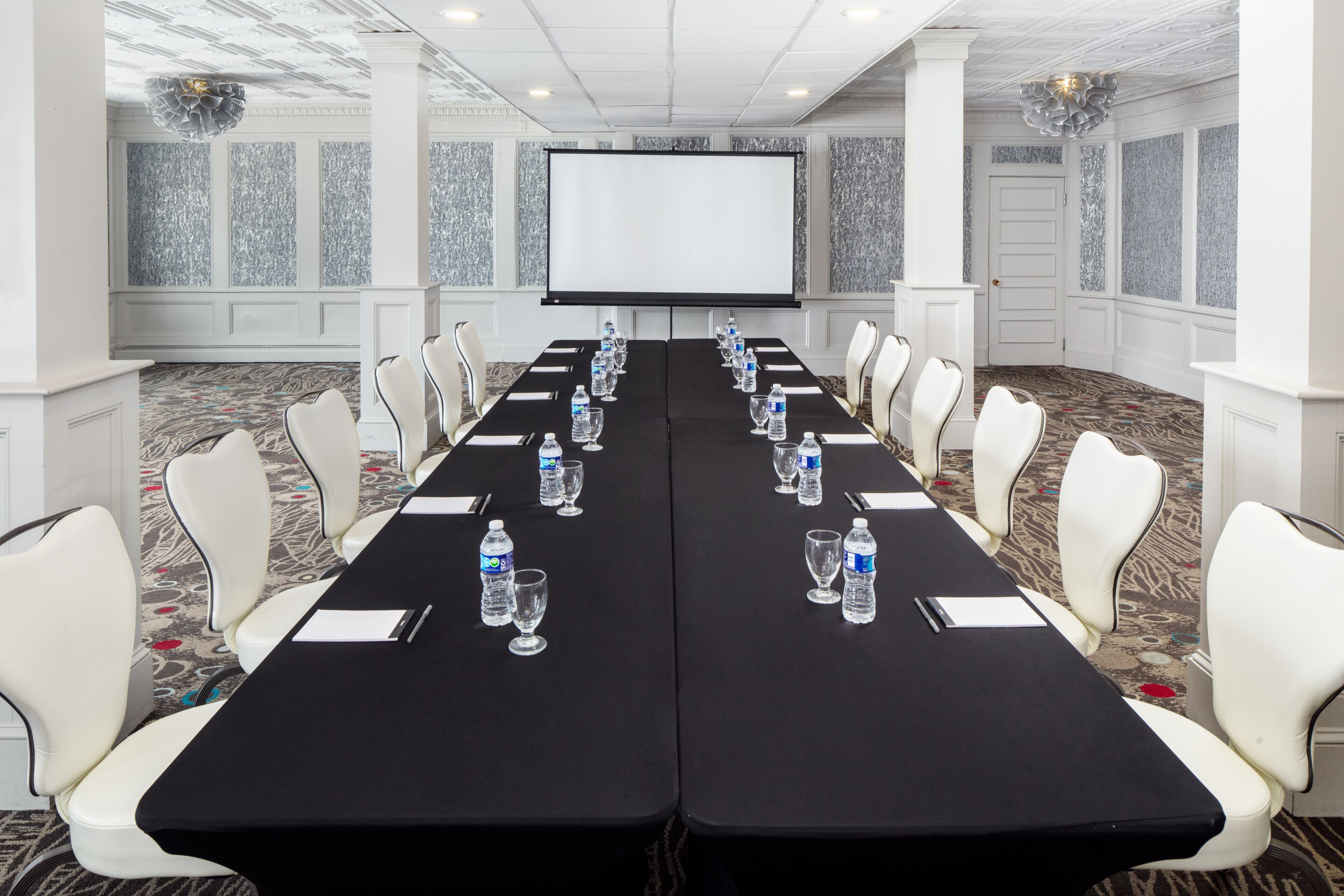 A conference room with a long table, white chairs, water bottles, and a projector screen.
