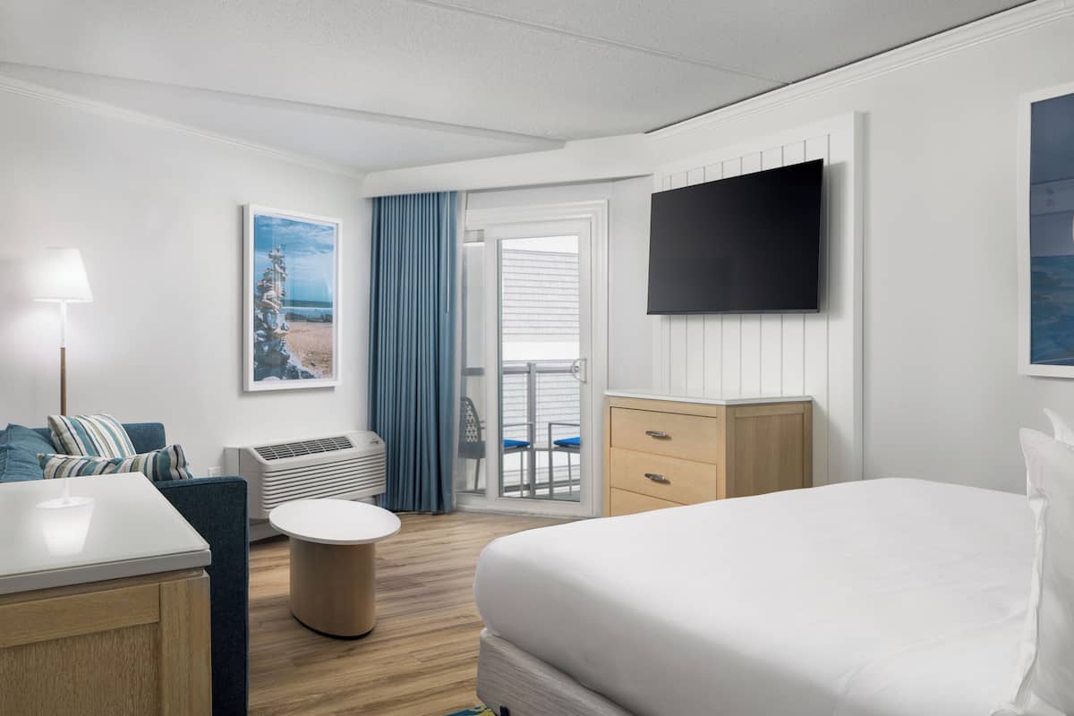 Ein Hotelzimmer mit Bett, Schreibtisch, Stuhl, Fernseher, einem Fenster mit Vorhängen und einer Tür, die auf einen Balkon führt.