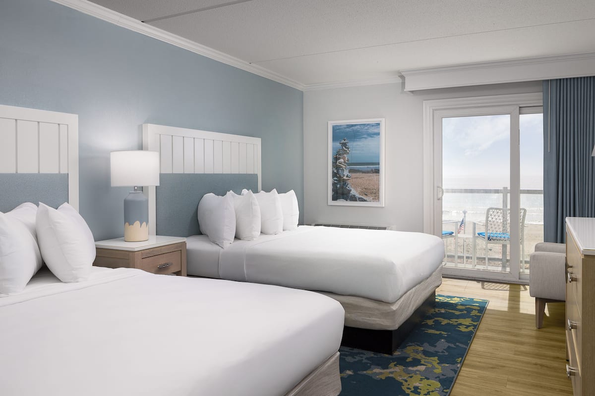 Ein Hotelzimmer mit zwei Betten, einem Nachttisch, einem Schreibtisch und Blick auf den Strand.
