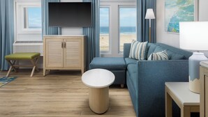 A modern living room with a large flat-screen TV, a sofa, a round coffee table, and a view of the beach. - Ashworth by the Sea (Hampton)