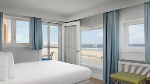 A hotel room with a bed, a bench, a view of the beach, and a window with curtains. - Ashworth by the Sea (Hampton)