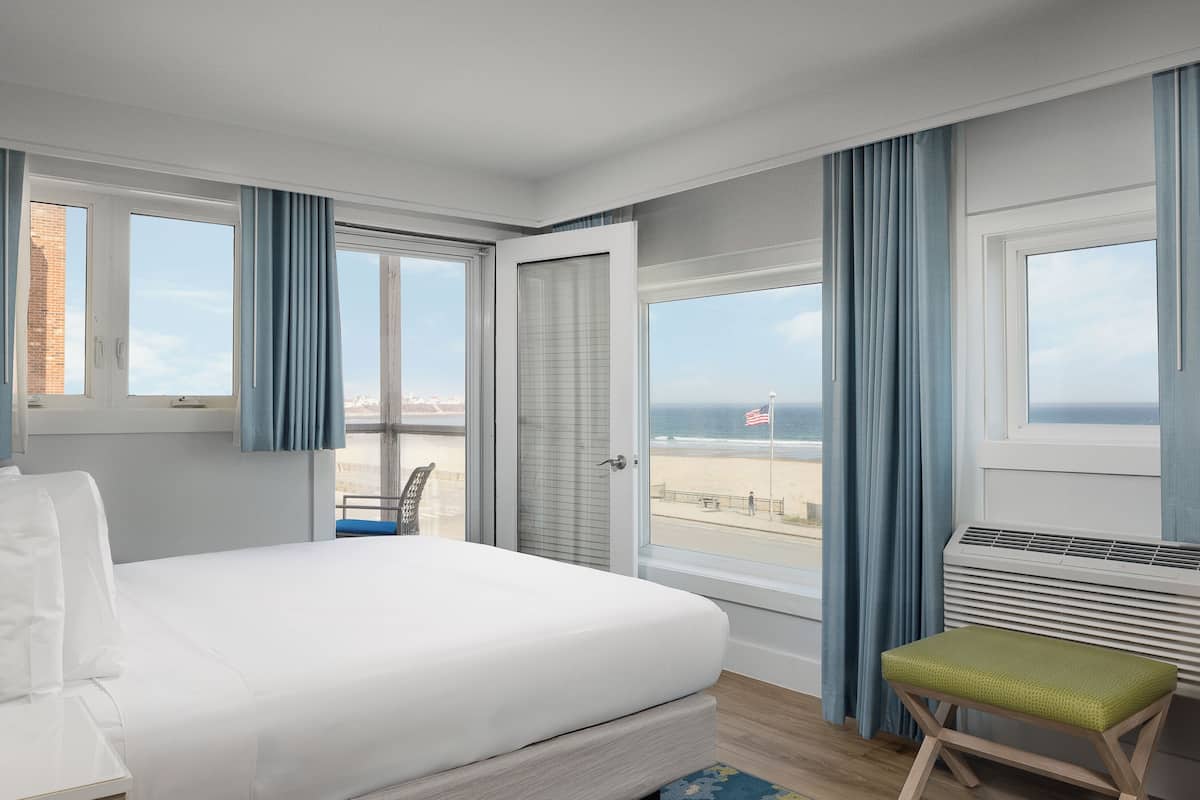 Ein Hotelzimmer mit Bett, Sitzbank, Blick auf den Strand und einem Vorhangfenster.