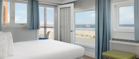 A hotel room with a bed, a bench, a view of the beach, and a window with curtains.