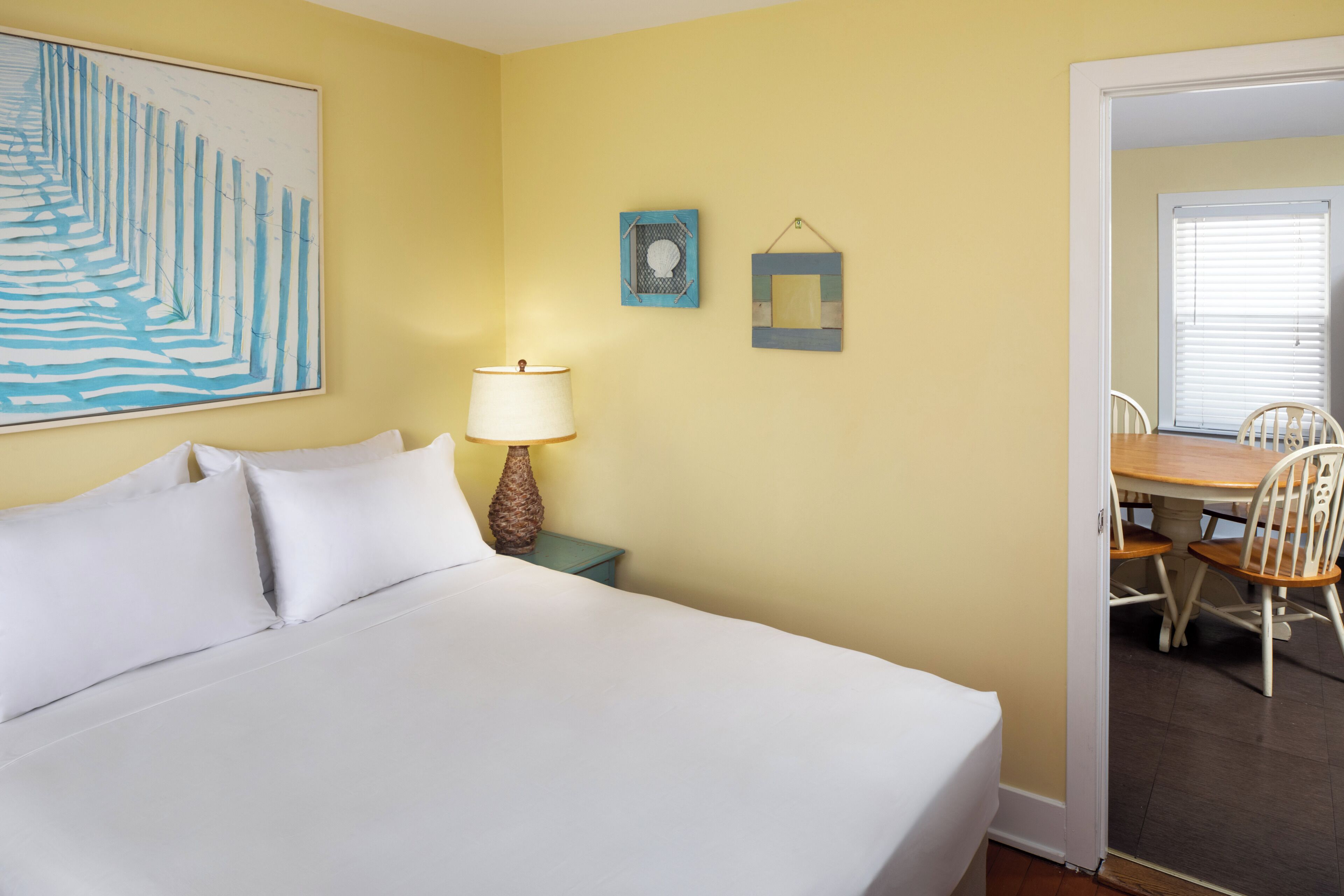 A hotel room with a bed, a nightstand with a lamp, a painting on the wall, and a dining area with chairs and a table.