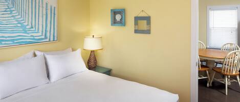 Quarto de hotel com uma cama, uma mesinha de cabeceira com abajur, um quadro na parede e um espaço de refeições com cadeiras e uma mesa.