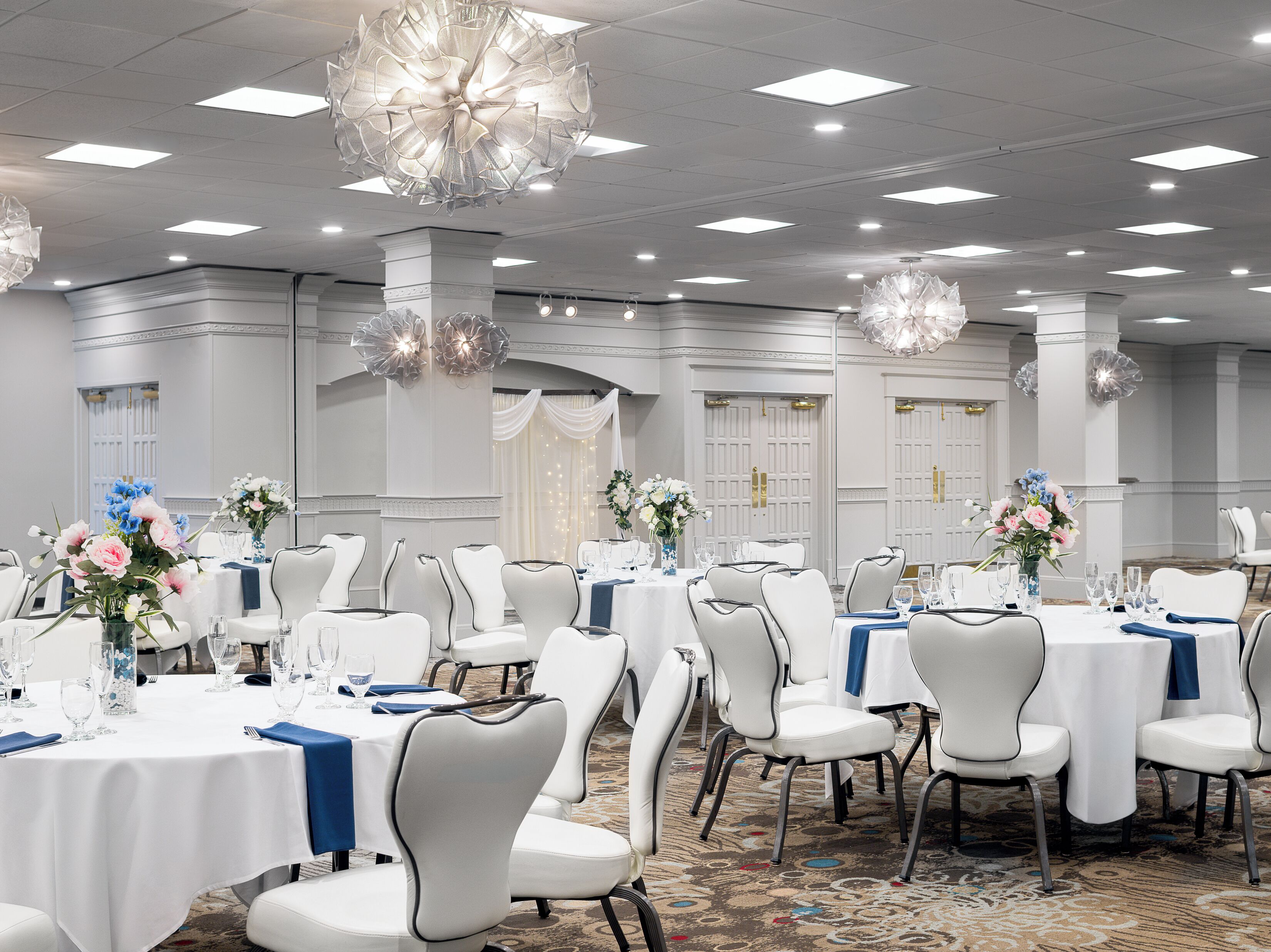 A banquet hall with round tables, white chairs, and chandeliers.