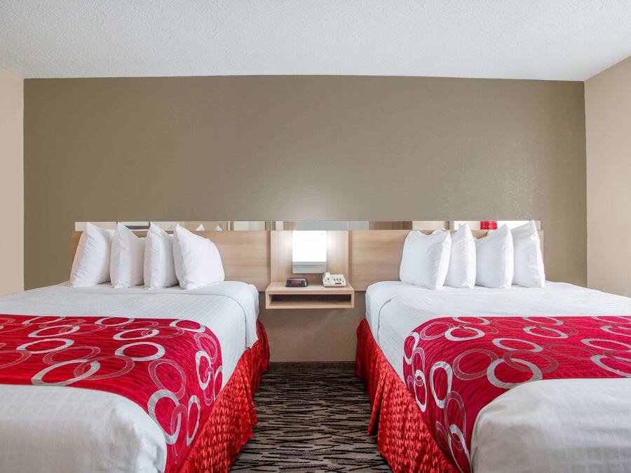 A hotel room with two beds, each with white bedding and red accents, a small bedside table with a lamp, and a mirror above the bed.