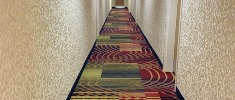 Un couloir d’hôtel avec un tapis à motifs, des murs de couleur crème et une porte ornée d’une enseigne bleue.