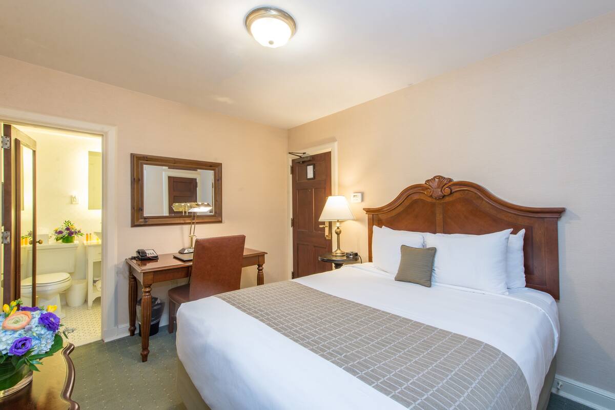 A hotel room with a large bed, a wooden desk, a chair, a mirror, and a bathroom visible through an open door.