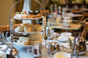 A three-tiered tea service stand with various pastries and sandwiches, surrounded by silver teapots and cups on a marble table. - Hotel deLuxe (Portland)