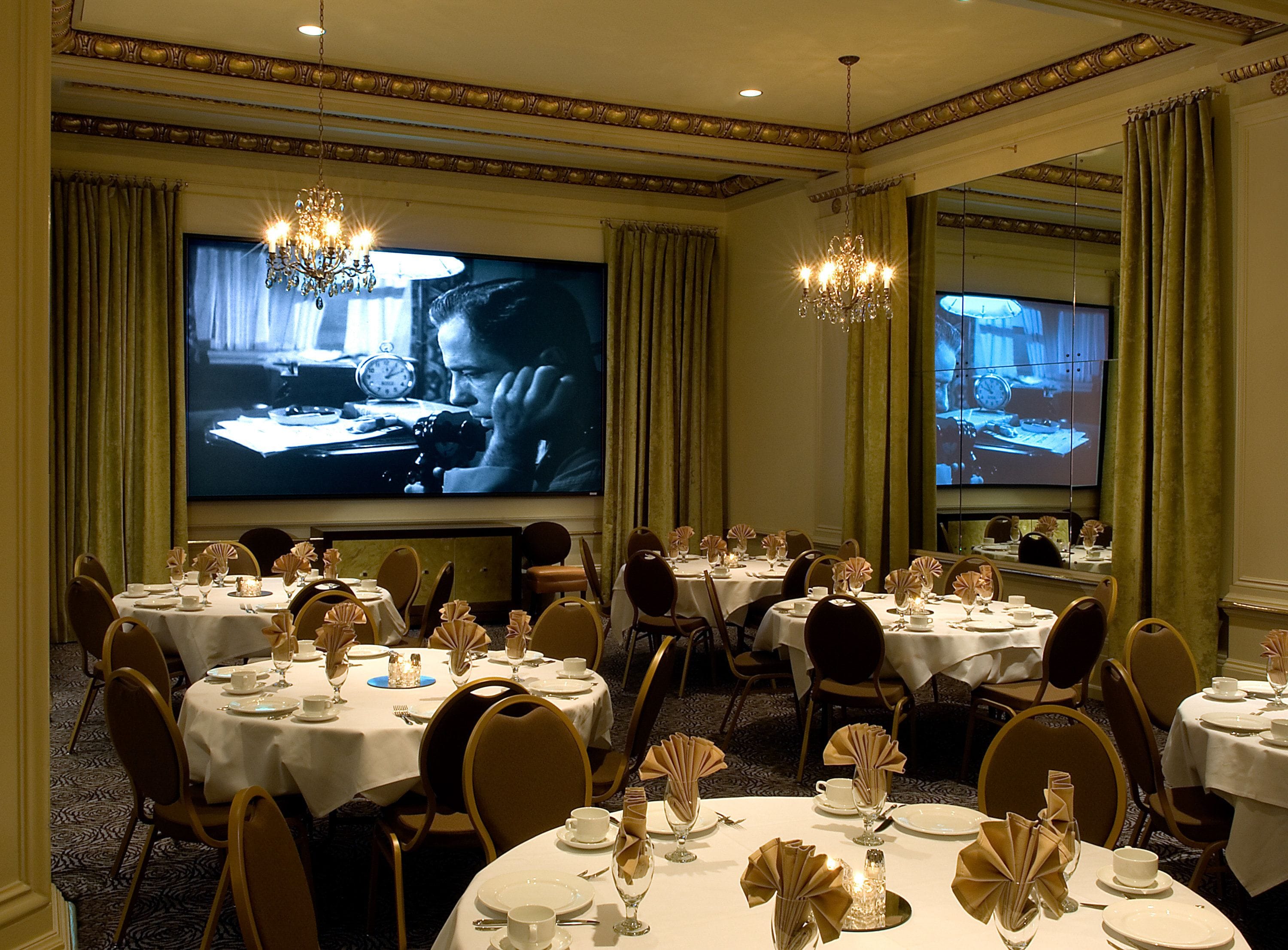A banquet hall with multiple round tables set for dining, a large screen displaying a program, and chandeliers.