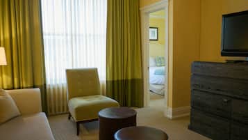 A hotel room with a sofa, armchair, two ottomans, a dresser, and a television.