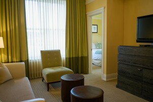 Habitación de hotel con sofá, sillón, dos otomanas, cómoda y televisor.