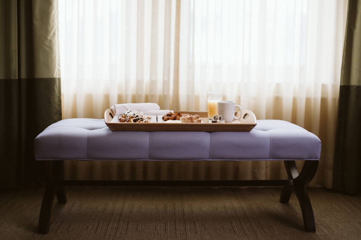 Habitación con un banco y una bandeja de desayuno, que incluye bollería y un vaso de zumo de naranja, cerca de una ventana con cortinas translúcidas.
