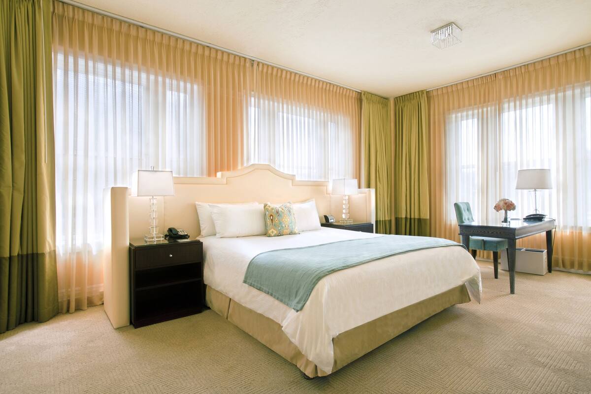 Una habitación de hotel con una cama grande, dos mesitas de noche con lámparas, un escritorio con silla y cortinas.