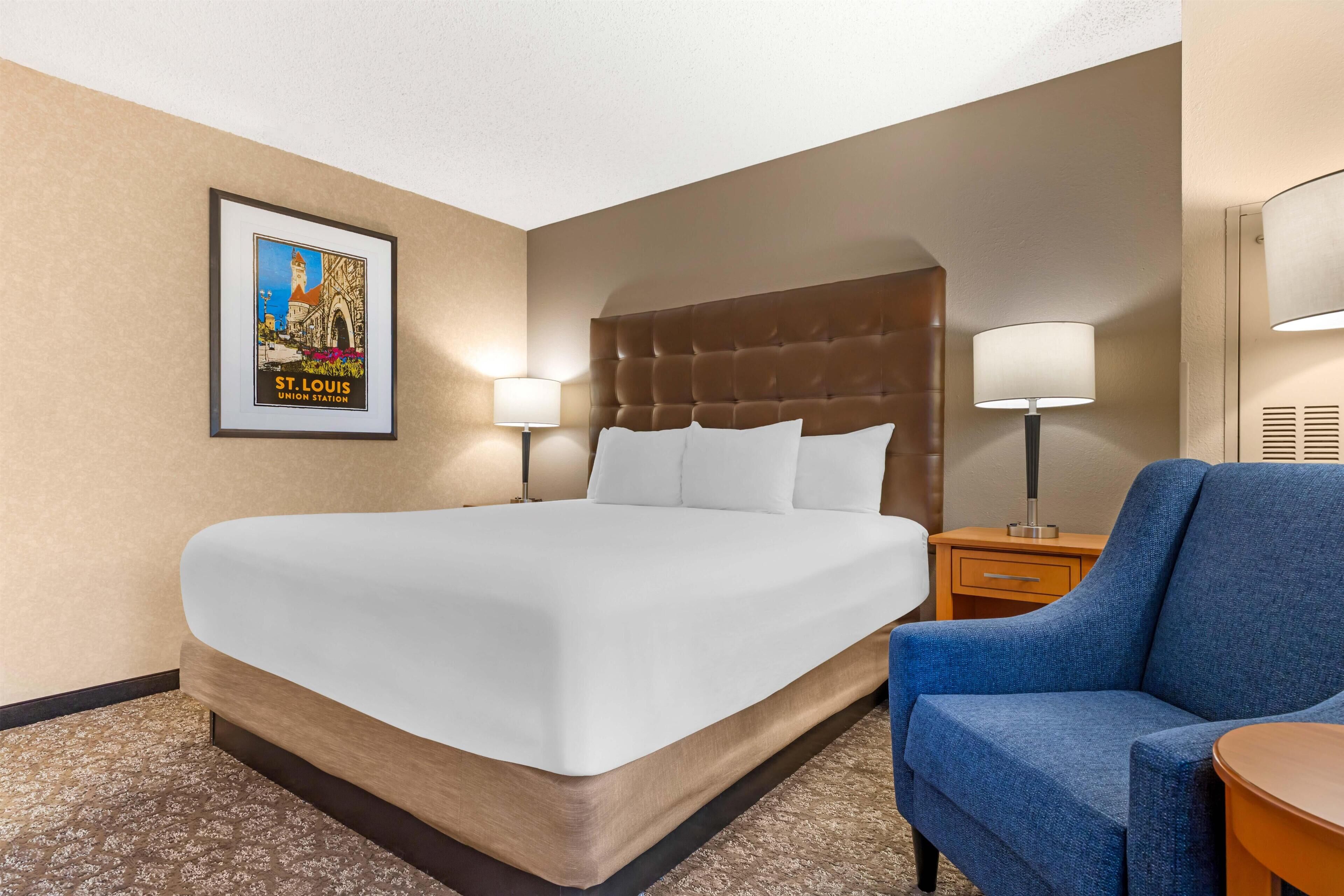 A hotel room with a large bed, two bedside lamps, a framed picture on the wall, and a blue armchair.