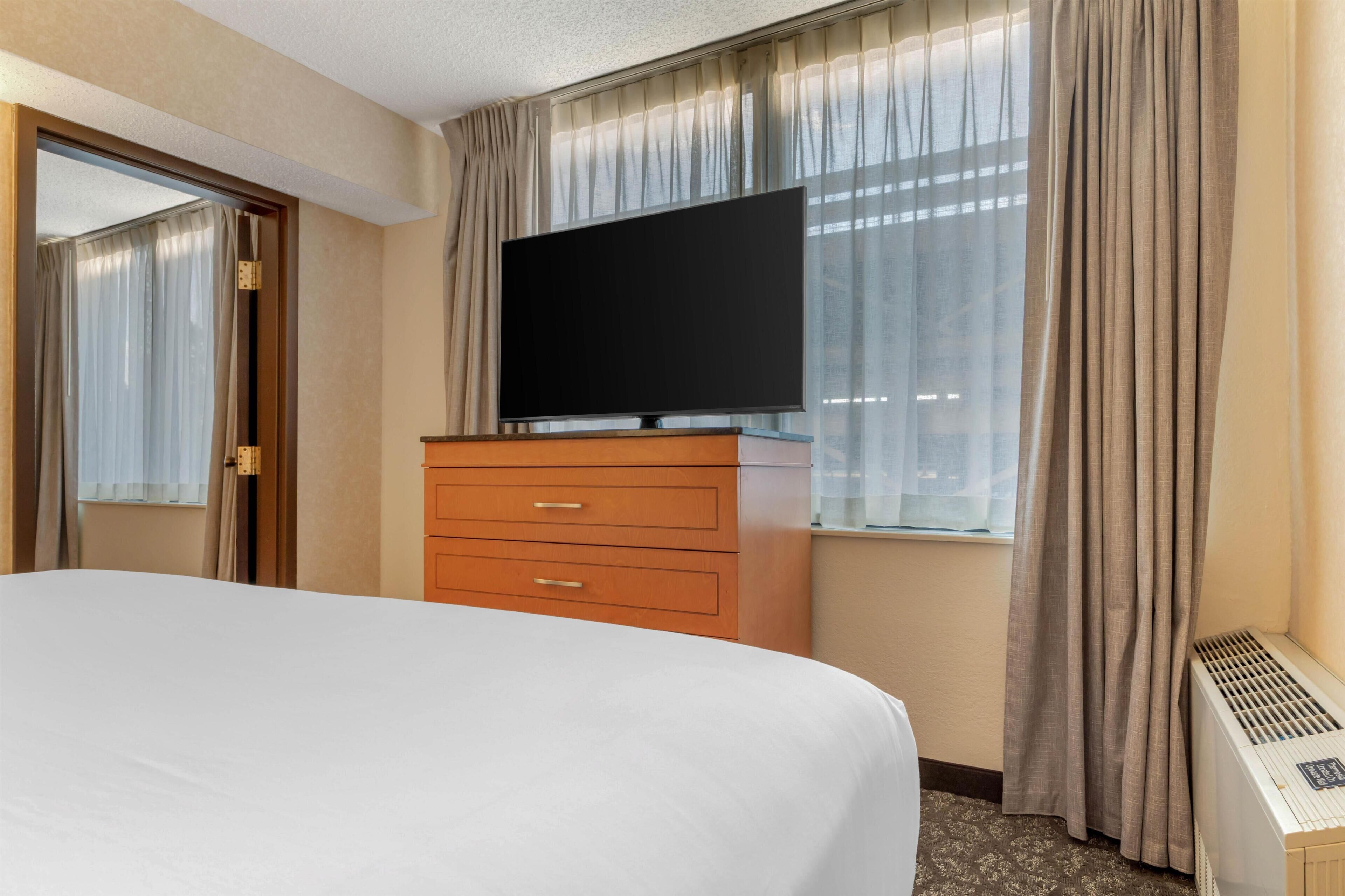 A hotel room with a bed, a TV on a wooden dresser, a window with curtains, and a heater.