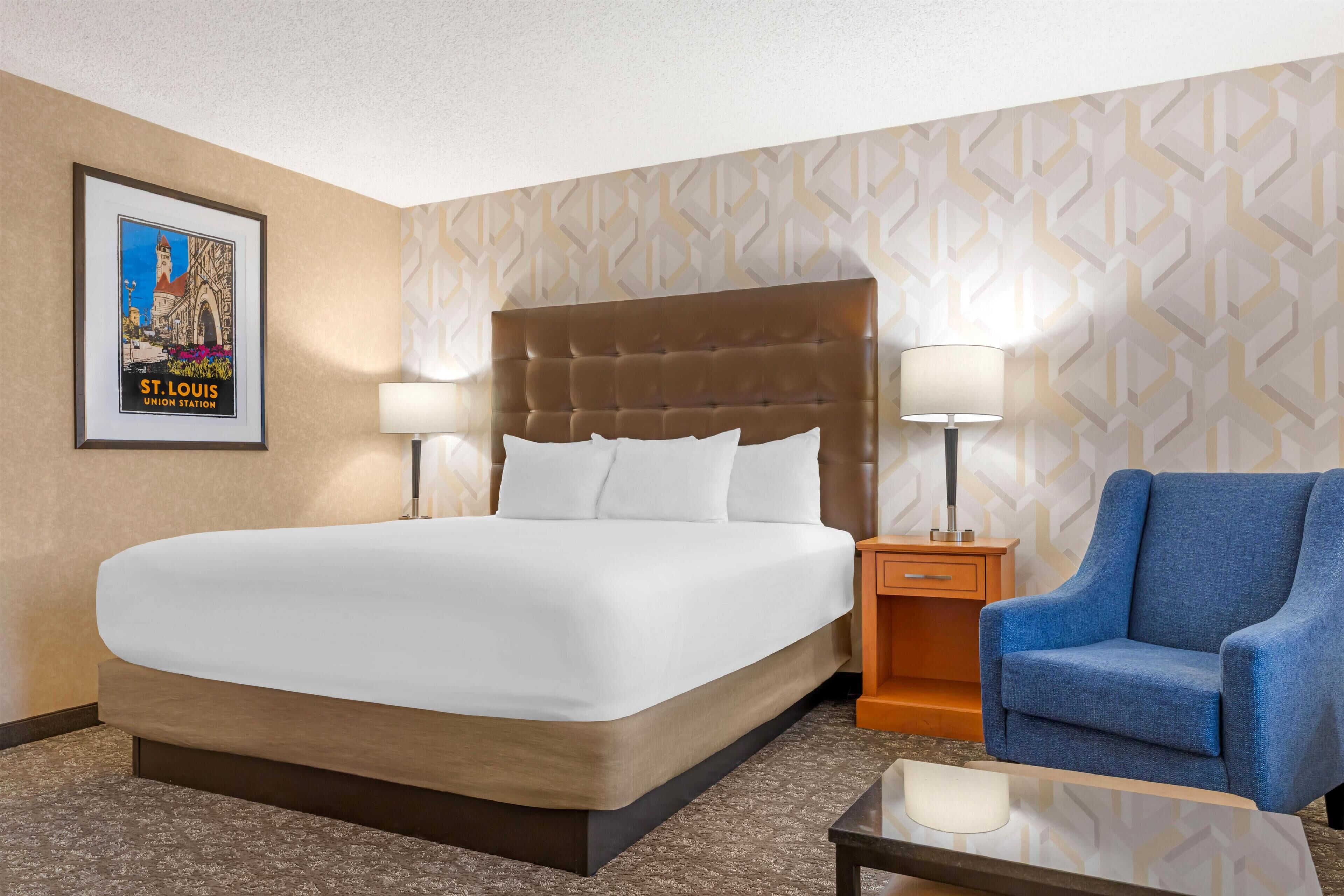 A hotel room with a large bed, two bedside lamps, a blue armchair, a small table, and a poster on the wall.