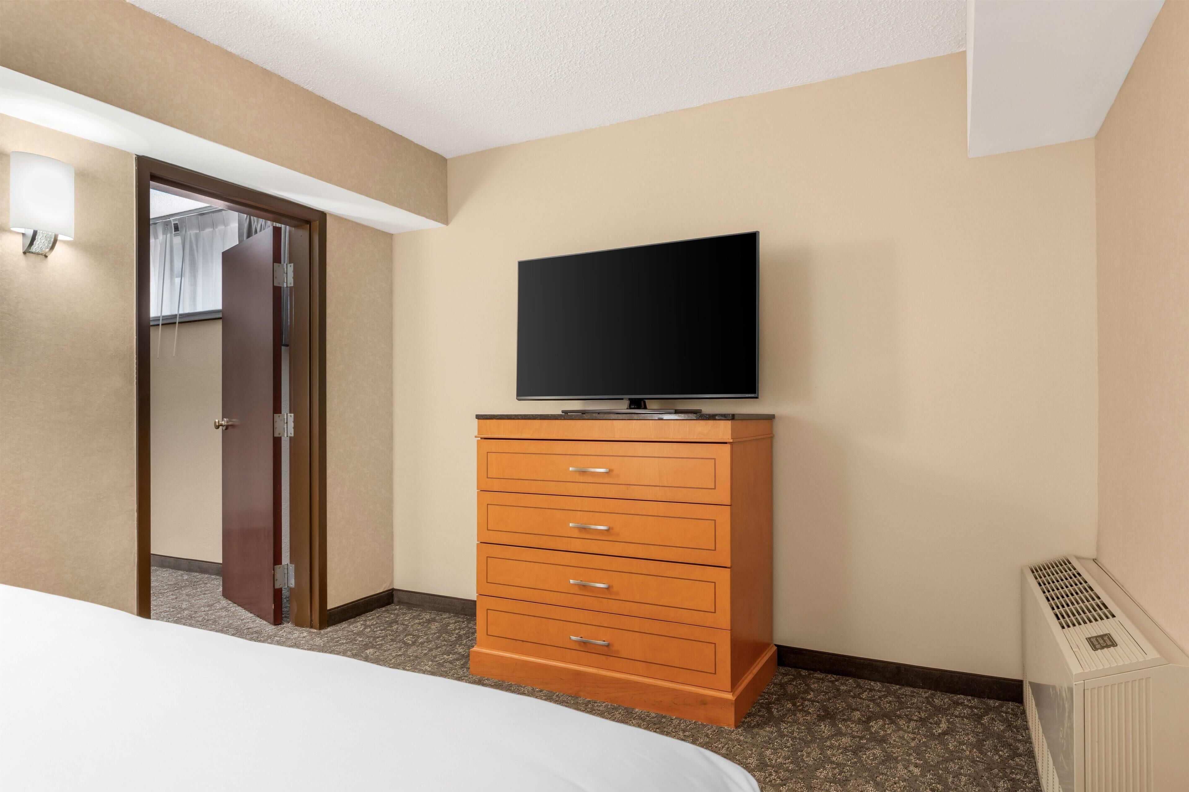 Une chambre d’hôtel avec un lit, une commode en bois dotée de tiroirs, un téléviseur posé sur le dessus, et une porte donnant sur une autre pièce.
