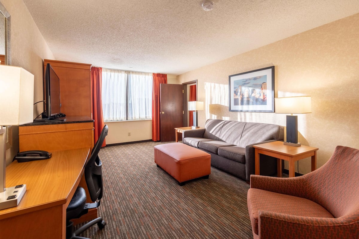 A hotel room with a desk, a sofa, an ottoman, a TV, a lamp, and a framed picture on the wall.