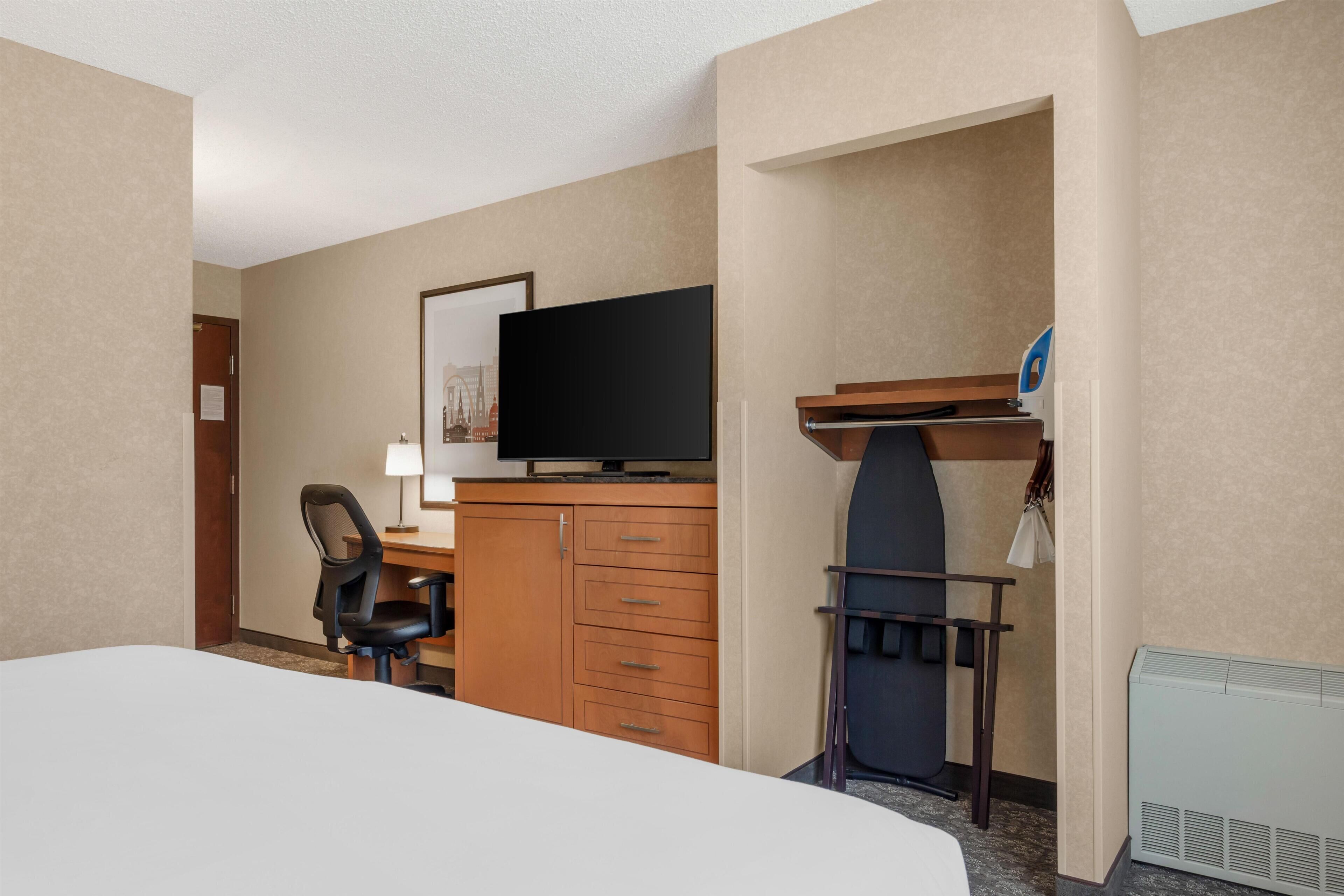 A hotel room with a bed, a desk with a flat-screen TV, a chair, a dresser, and a coat rack.