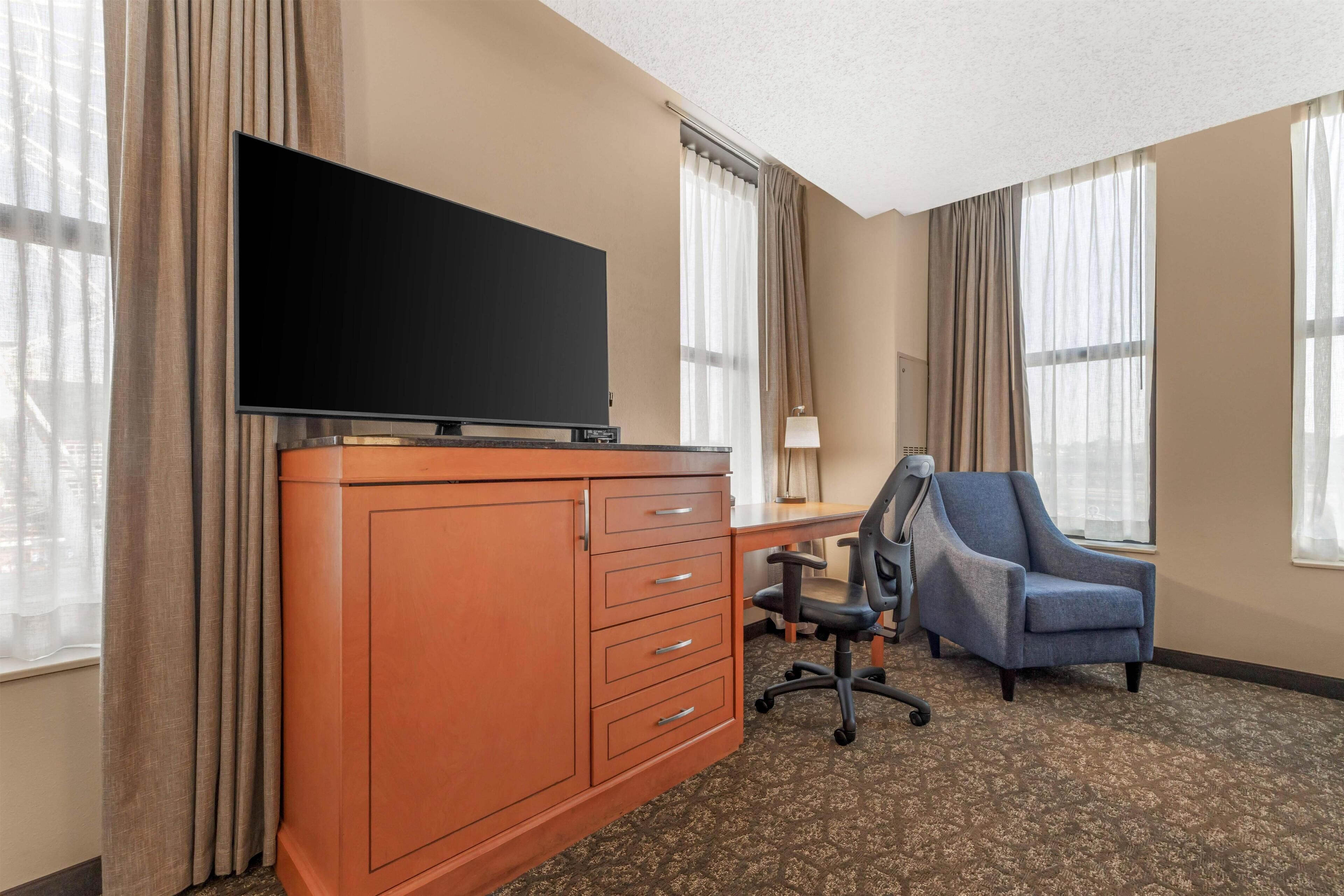 Ein Hotelzimmer mit einem Flachbildfernseher auf einer Holzkommode, einem Schreibtisch mit Stuhl und großen Fenstern mit Vorhängen.