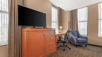 Ein Hotelzimmer mit einem Flachbildfernseher auf einer Holzkommode, einem Schreibtisch mit Stuhl und großen Fenstern mit Vorhängen.