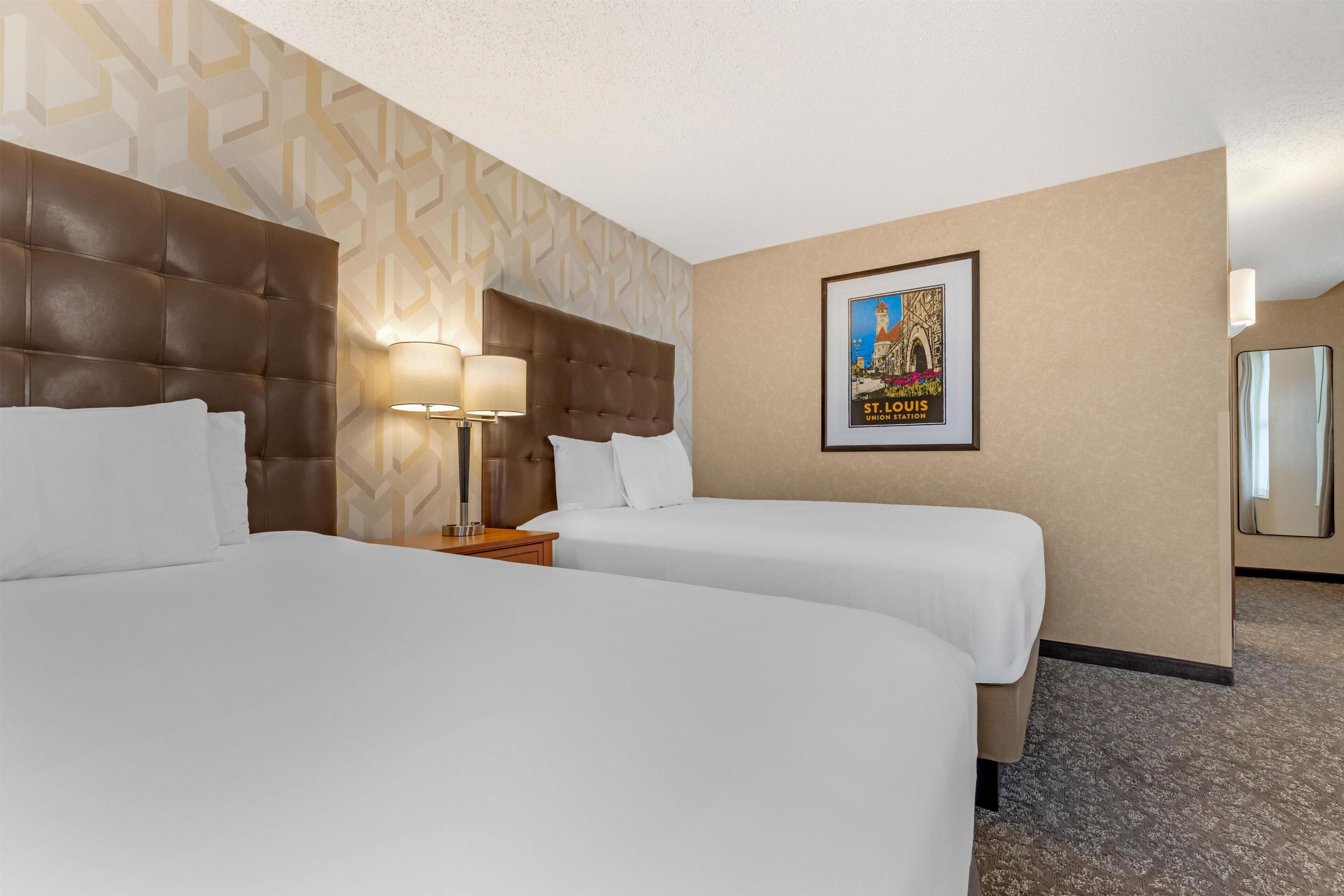 A hotel room with two beds, a headboard, a nightstand with lamps, a framed picture on the wall, and a carpeted floor.