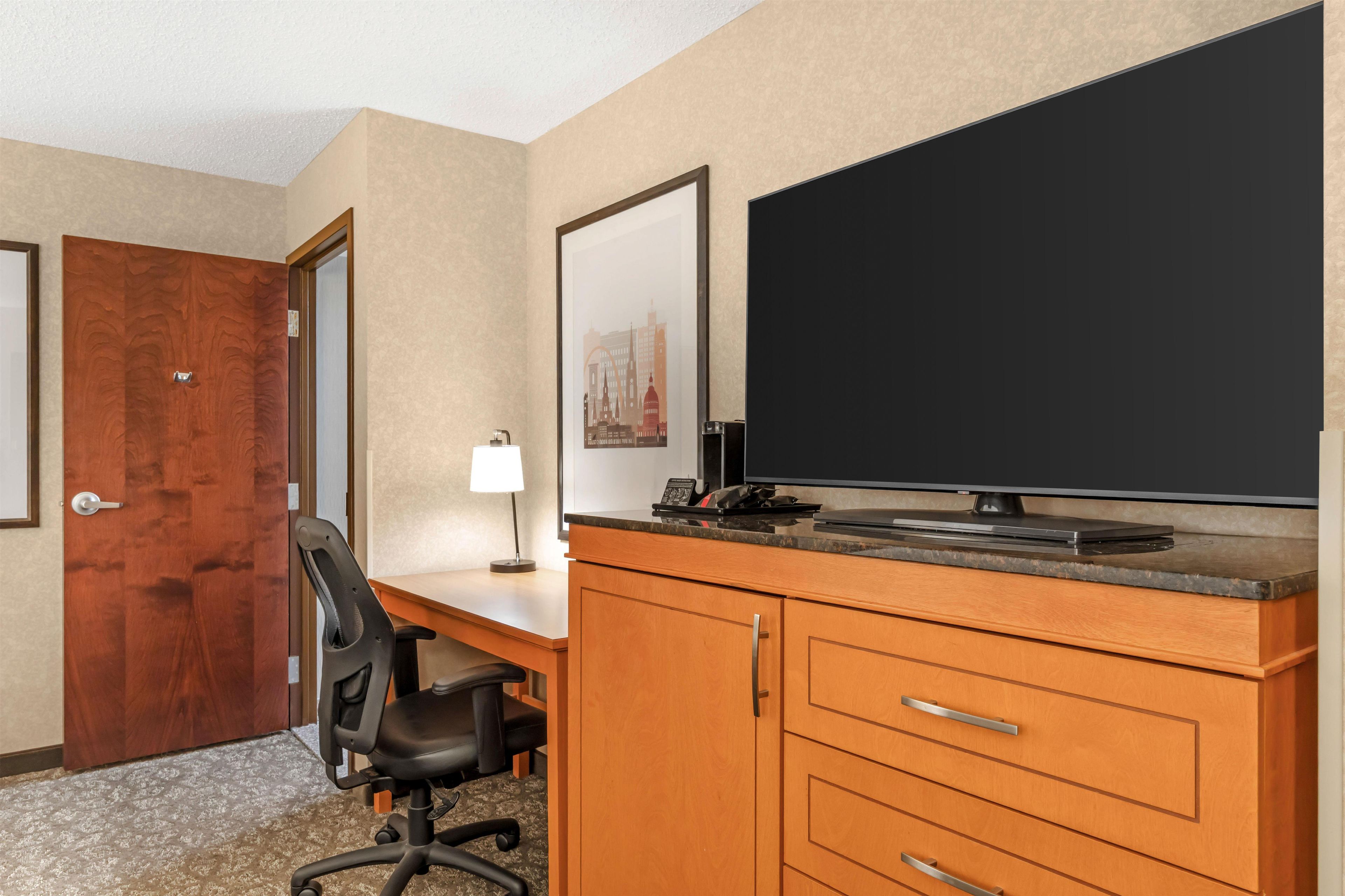 Quarto de hotel com escrivaninha de madeira, cadeira de escritório preta, televisão de tela plana, luminária e porta.